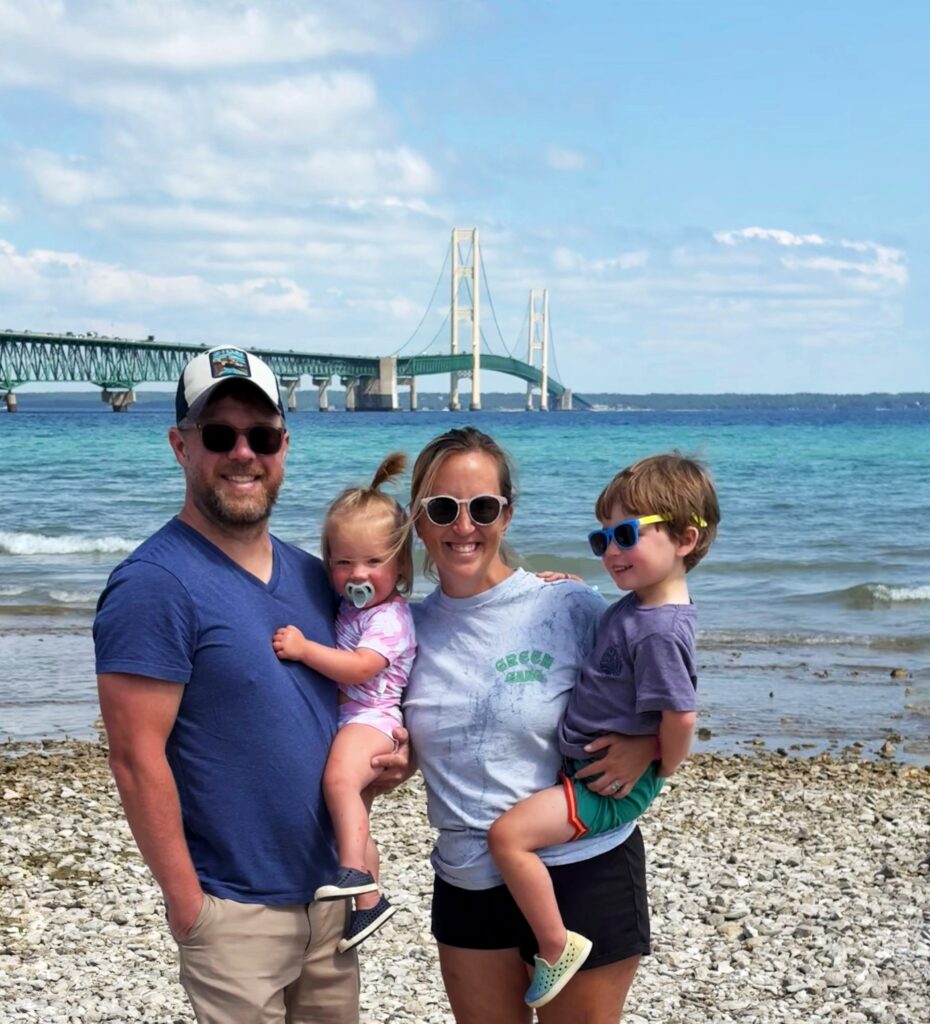 A smiling family of four stands on a rocky beach in front of a large bridge over blue water. The adults hold two young children, all dressed in summer clothes and sunglasses, with a bright sky overhead.