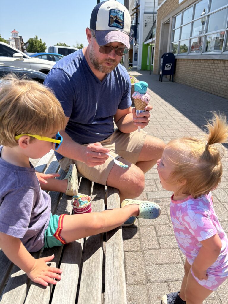 A man in a blue shirt and cap holds ice cream while helping feed a little girl with a spoon. A boy in sunglasses sits on a bench with his own ice cream. All three are outside on a sunny day.