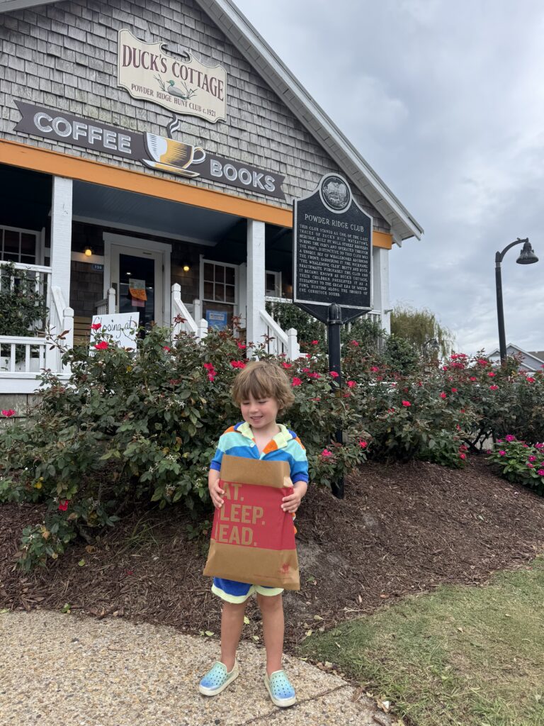 A young child stands smiling in front of Duck’s Cottage coffee and book shop, holding a brown bag that says “KEEP. READ.” Pink flowers and a historic sign are visible in the background.