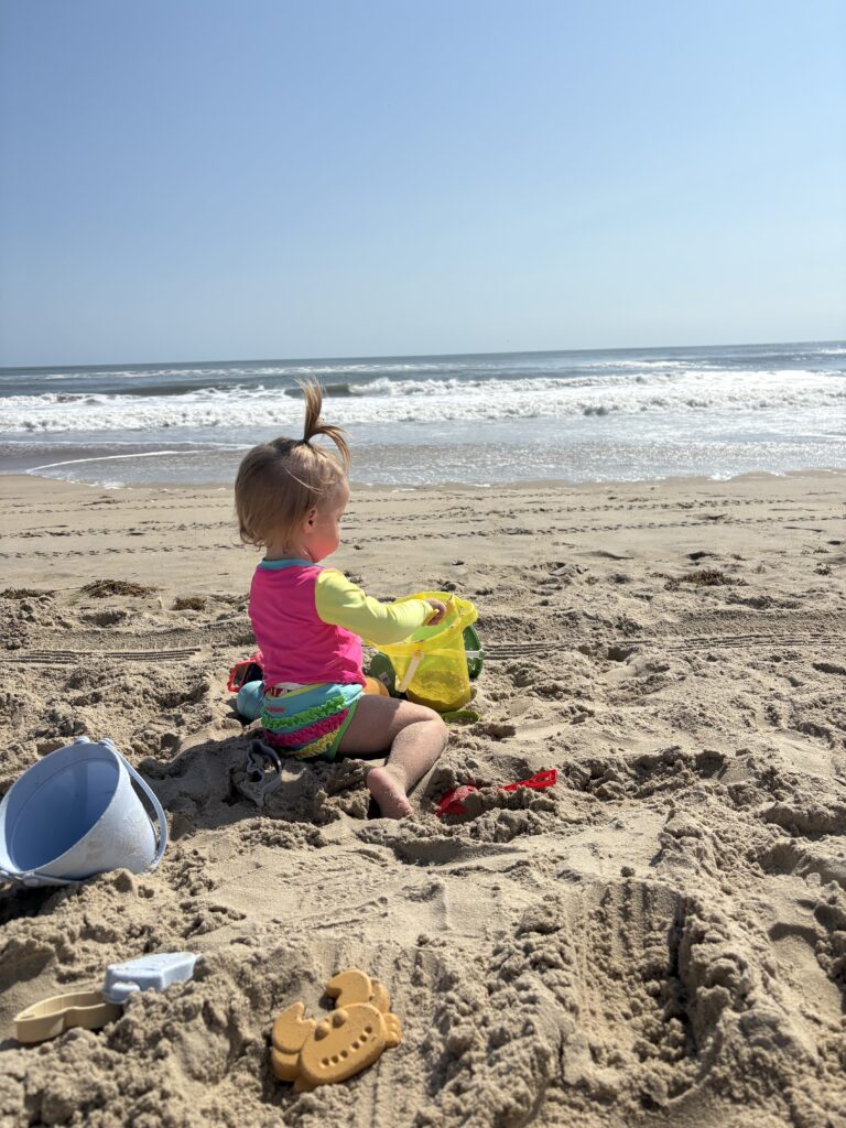 A young child in colorful swimwear sits on sandy beach near the ocean, playing with a yellow bucket and surrounded by sand toys under a clear, sunny sky.