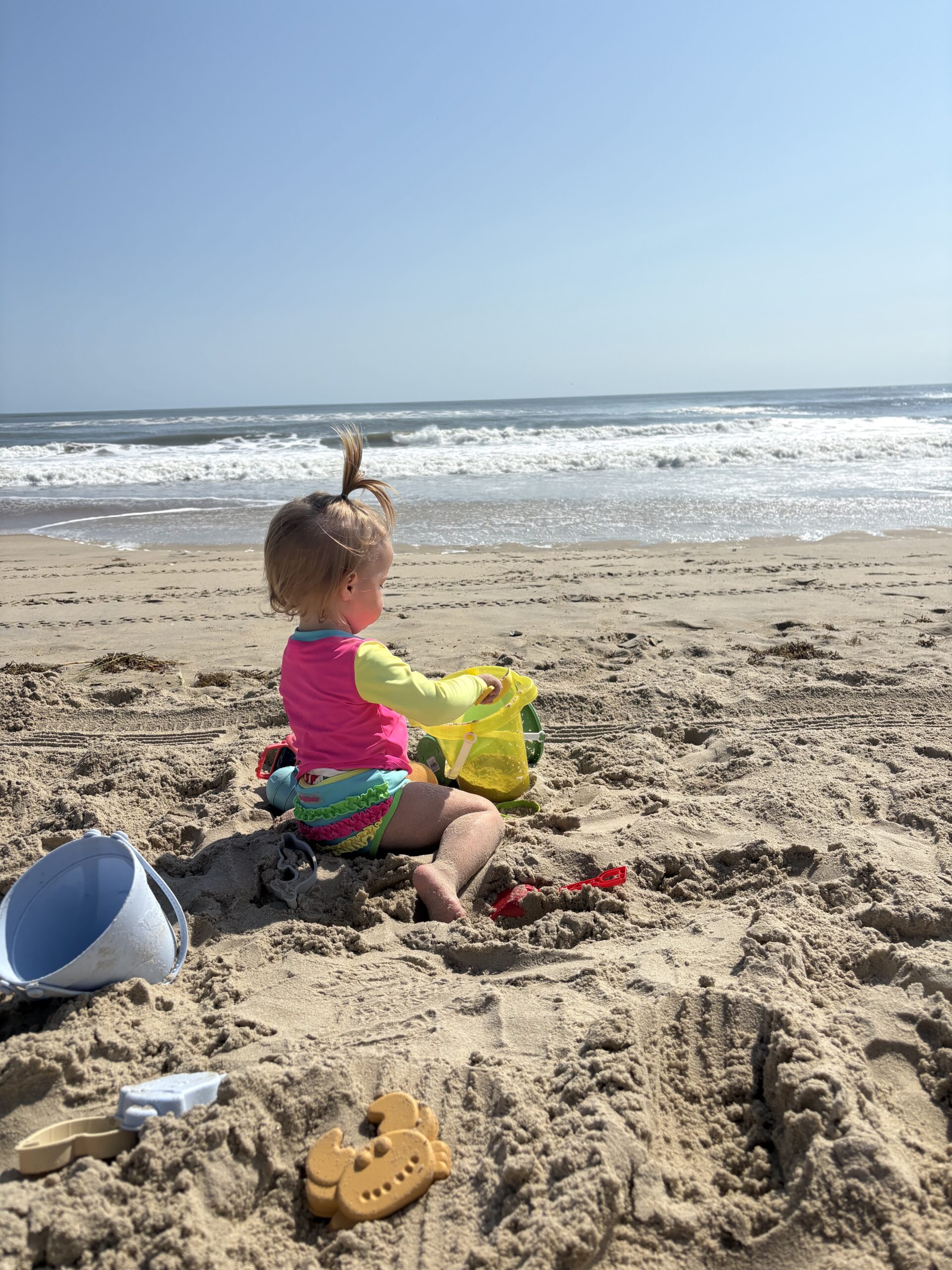A young child in colorful swimwear sits on sandy beach near the ocean, playing with a yellow bucket and surrounded by sand toys under a clear, sunny sky.