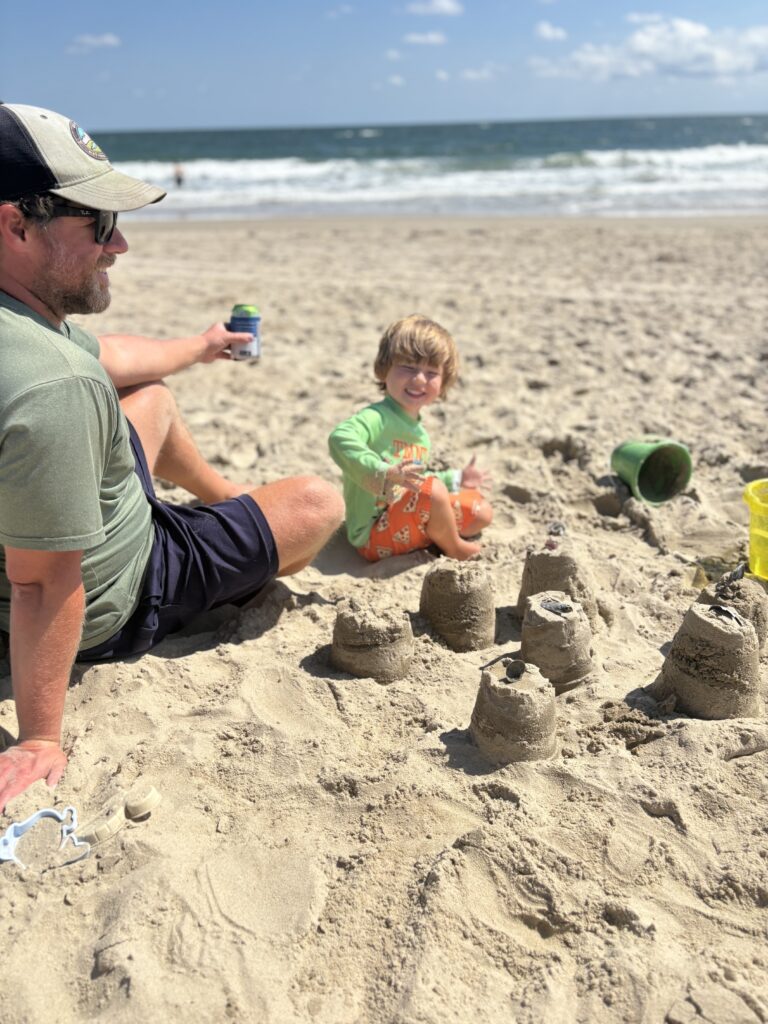 A man and a child sit on the sandy beach near several small sandcastles. The child smiles, wearing a green shirt and orange shorts, while the man holds a drink. The ocean and blue sky are in the background.