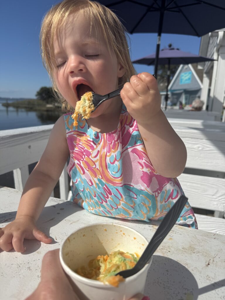 A young child in a colorful dress is sitting at an outdoor table, eagerly eating food from a cup with a black plastic fork. The child has eyes closed and mouth open wide in the sunshine.