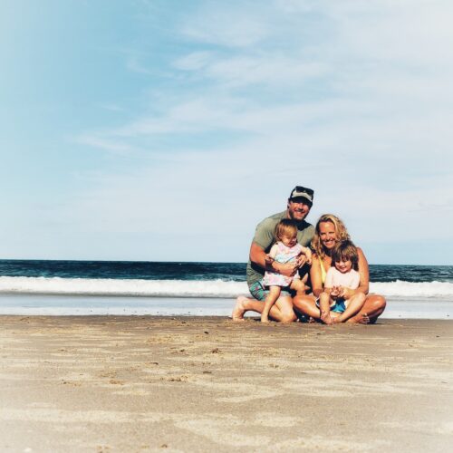 A family of four, consisting of two adults and two young children, kneels together on a sandy beach with waves and a blue sky in the background. They are smiling and appear happy.