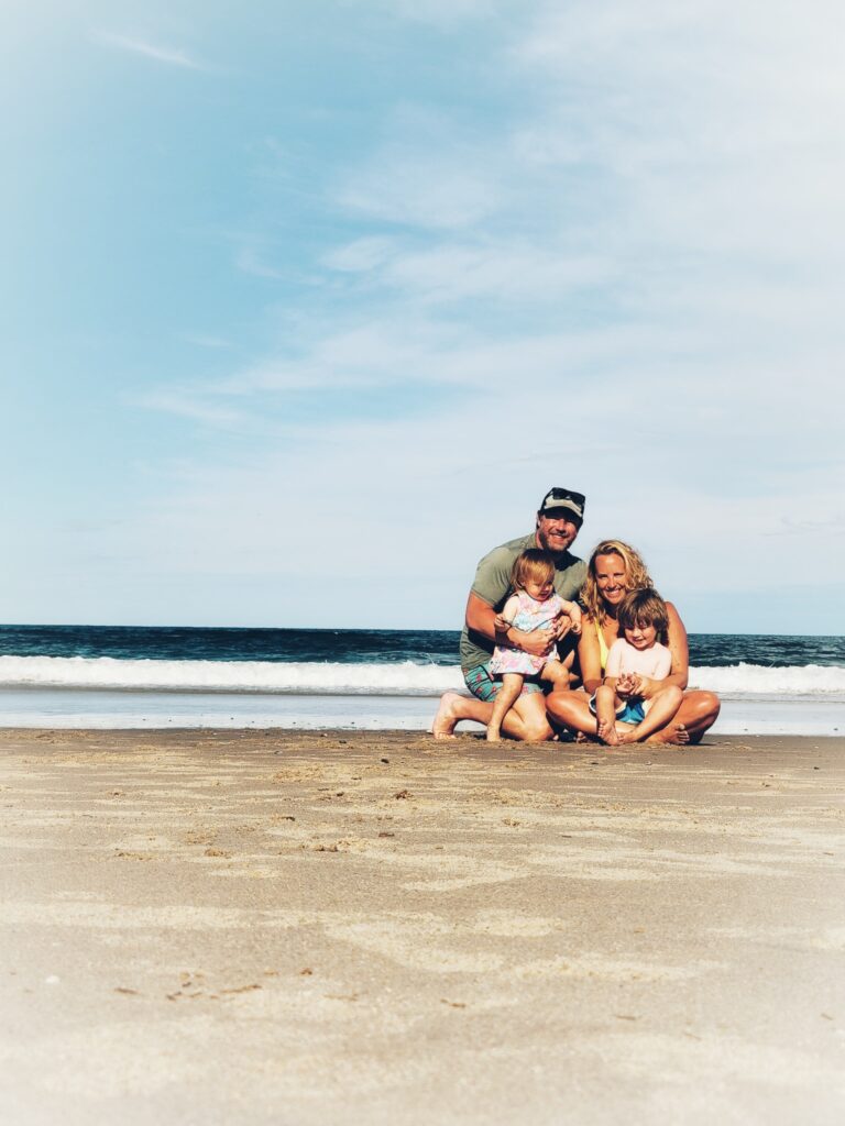 A family of four, consisting of two adults and two young children, kneels together on a sandy beach with waves and a blue sky in the background. They are smiling and appear happy.