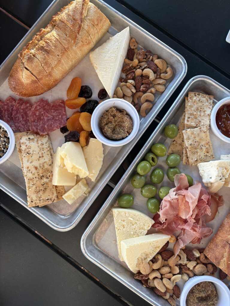 Two trays with assorted charcuterie including bread, different cheeses, crackers, nuts, green olives, dried fruits, prosciutto, salami, whole grain mustard, and a small jar of jam, all arranged neatly on parchment paper.