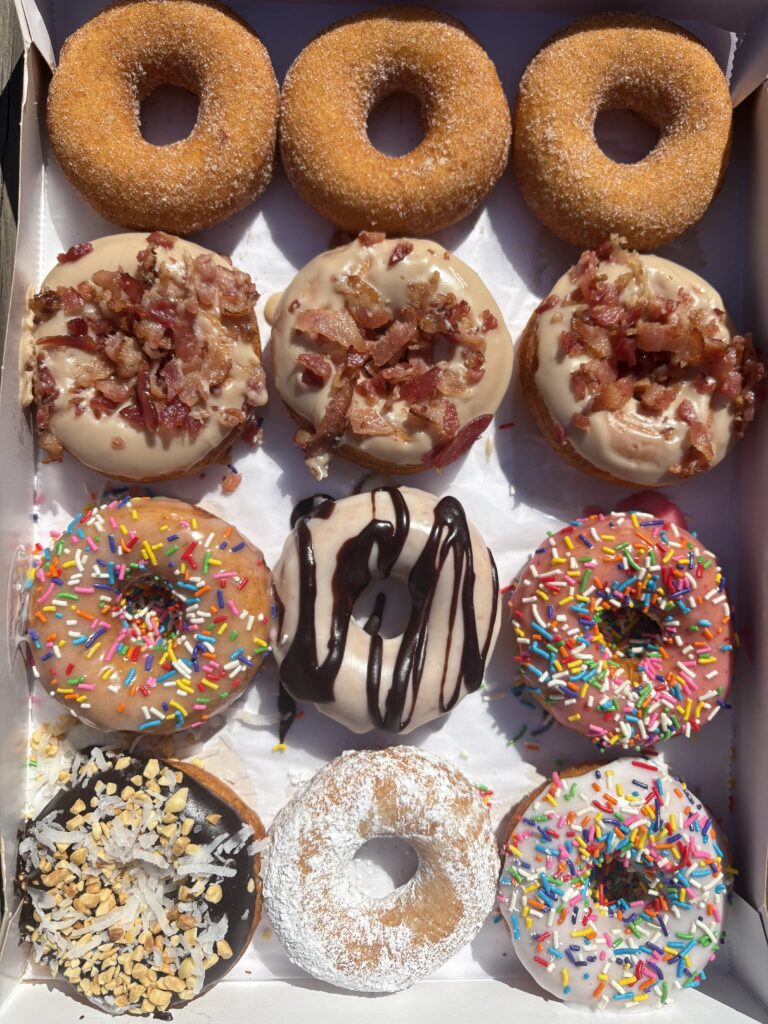 A dozen assorted donuts in a box, including plain sugared, maple bacon, rainbow-sprinkled, chocolate with sprinkles, powdered sugar, chocolate drizzle, and a coconut-topped donut, all brightly lit by sunlight.