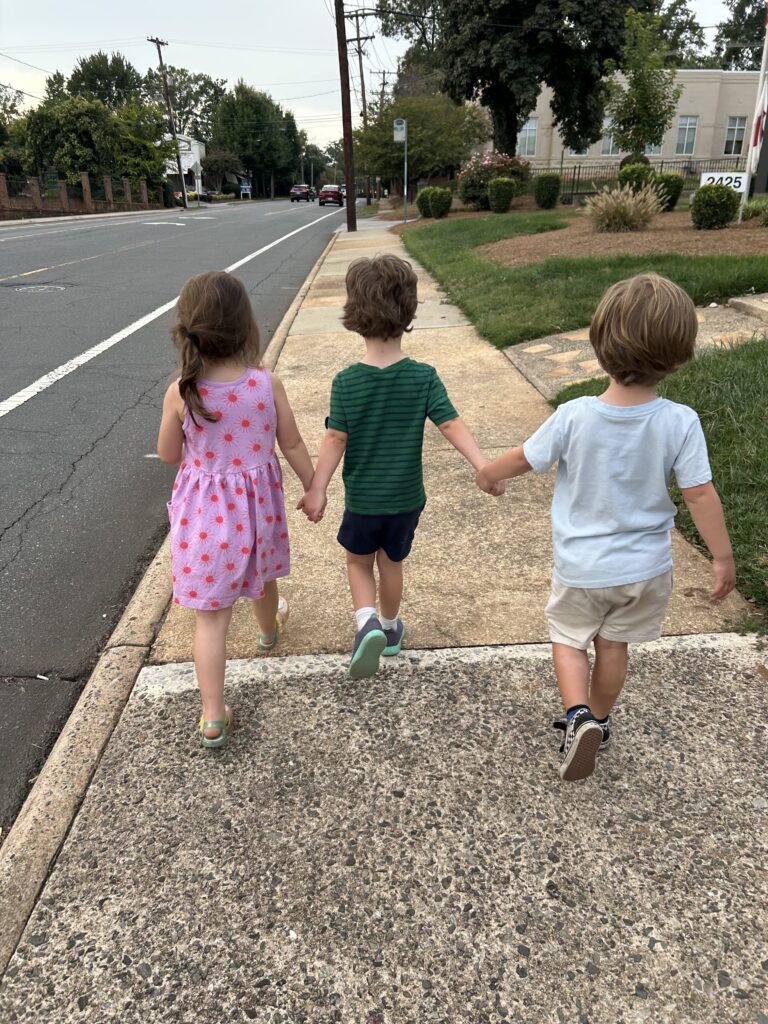 Three young children walk hand in hand on a sidewalk, heading away from the camera—just one of many sweet moments you’ll find exploring things to do in Charlotte with kids. Trees and houses line the street beside them.
