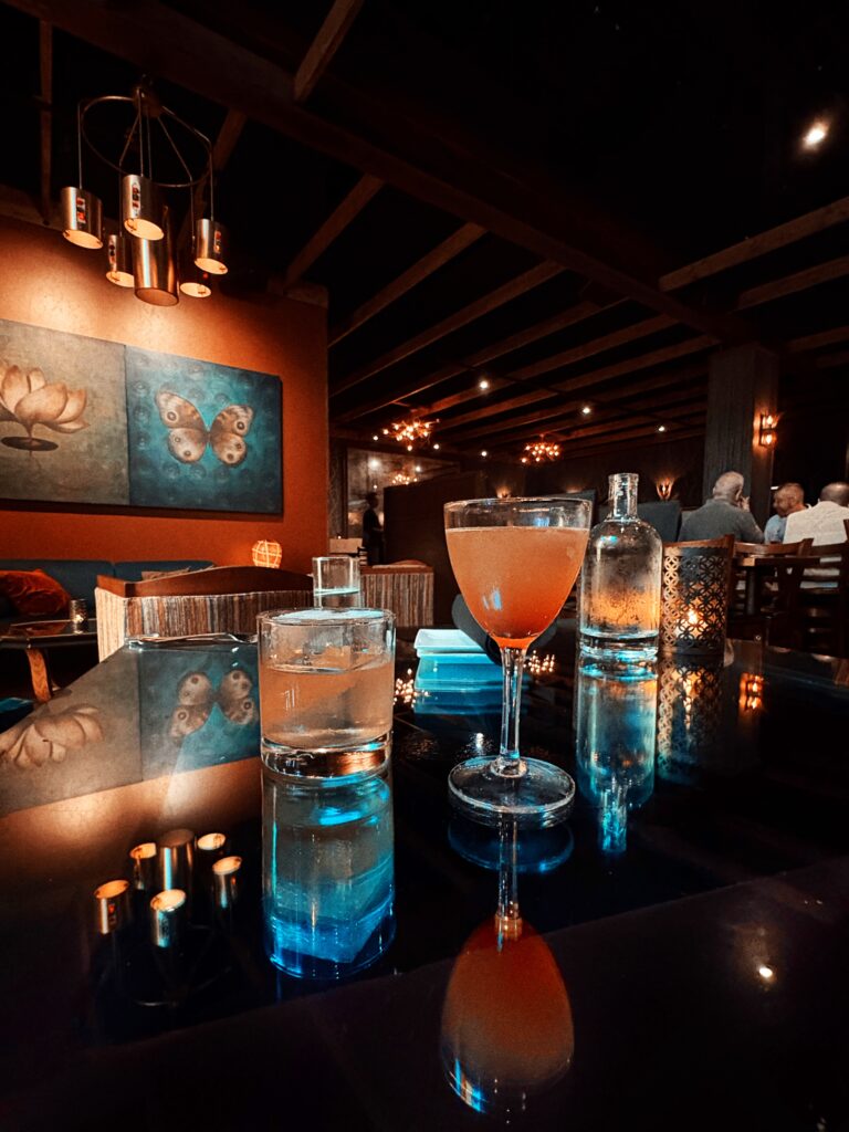 A dimly lit, elegant restaurant with two drinks on a reflective table—one in a rocks glass with ice, the other in a cocktail glass. Art and warm lighting decorate the cozy, intimate space.