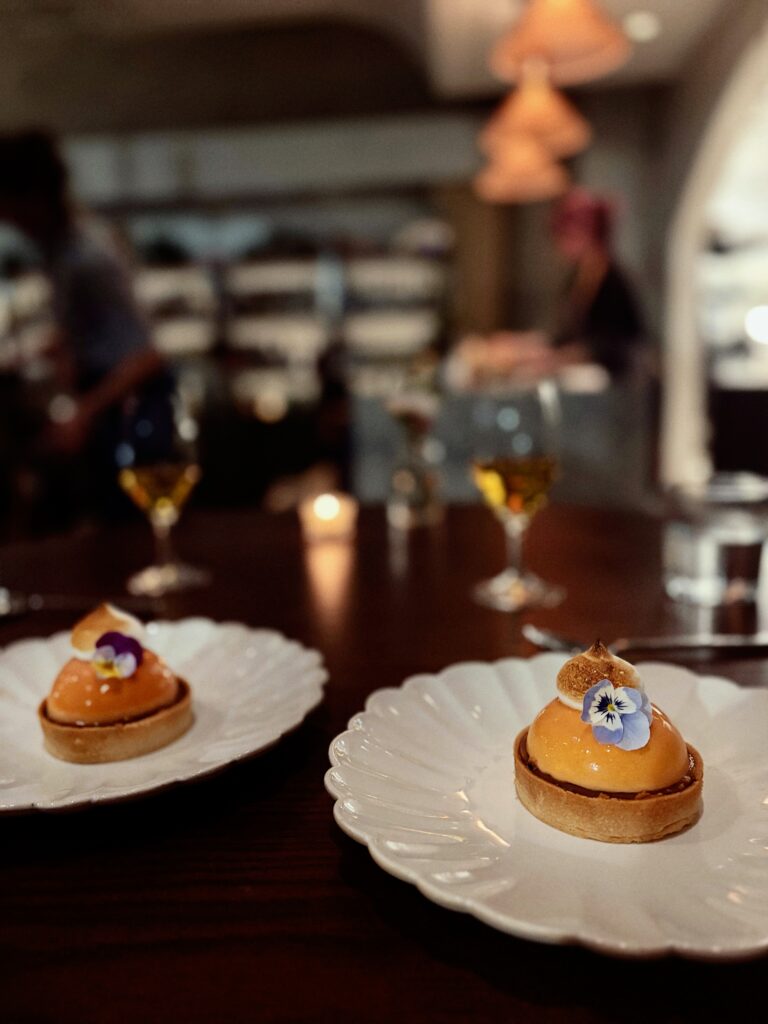 Two elegant pastries topped with orange glaze and edible flowers are served on white plates at a dimly lit restaurant table, with wine glasses and blurred diners in the background.