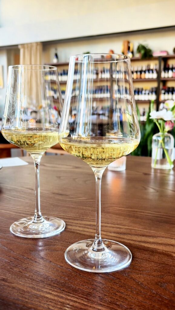 Two glasses of white wine sit on a wooden table in a bright, modern wine bar, with shelves of bottles and a vase with flowers blurred in the background.