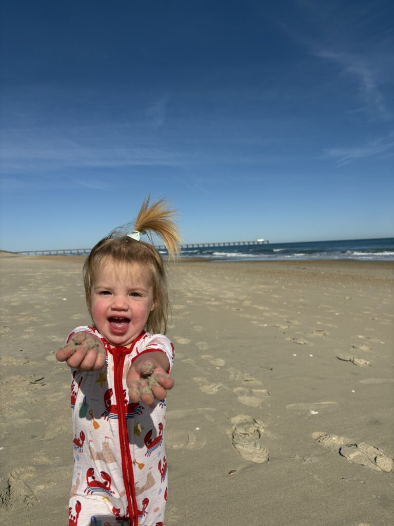 A smiling toddler in a lobster-printed outfit stands on a sandy beach, holding out sandy hands toward the camera. The sky is clear and blue, with a long pier and ocean waves visible in the background.