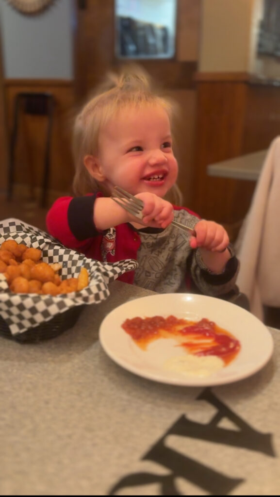 A smiling toddler holding two forks sits at a table with a basket of tater tots and a plate with ketchup and sauce. The child looks happy and is wearing a gray and red outfit.