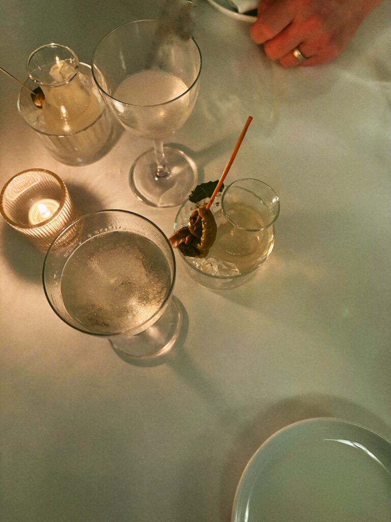 A tabletop with a candle, two filled wine glasses, two small carafes—one with a garnish—and a white plate. A hand wearing a ring rests at the top right corner of the image.