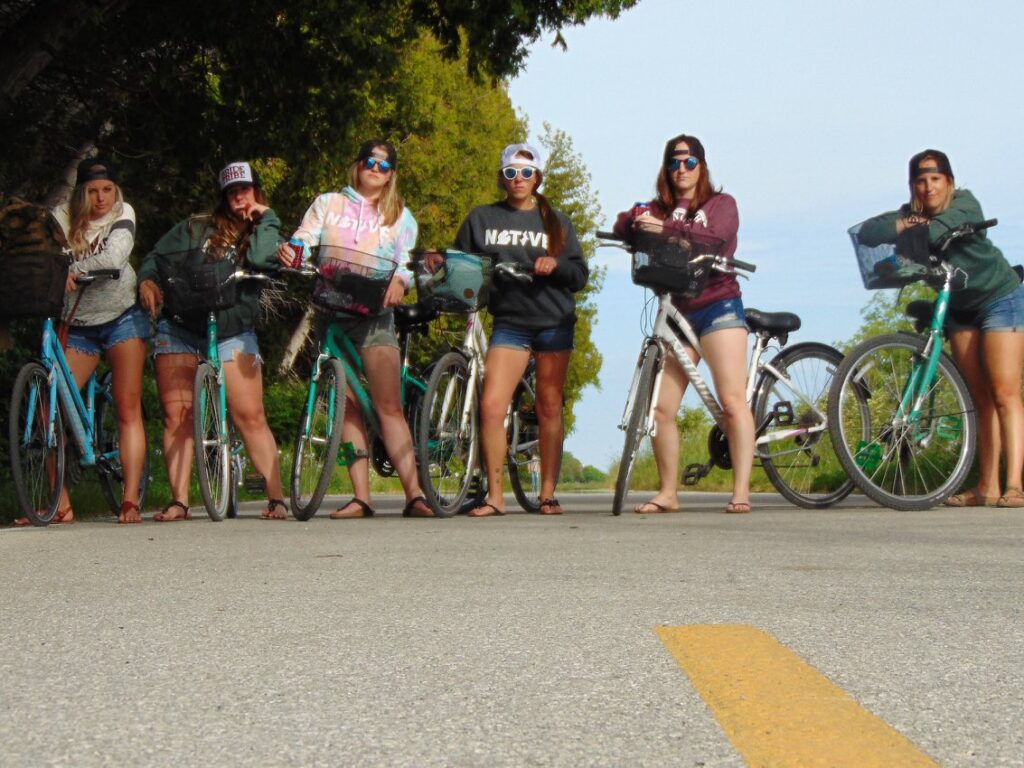 Six young women stand beside their bicycles in the middle of a road, posing for the photo. They are dressed casually in shorts, sweatshirts, and sunglasses, with trees and greenery in the background.