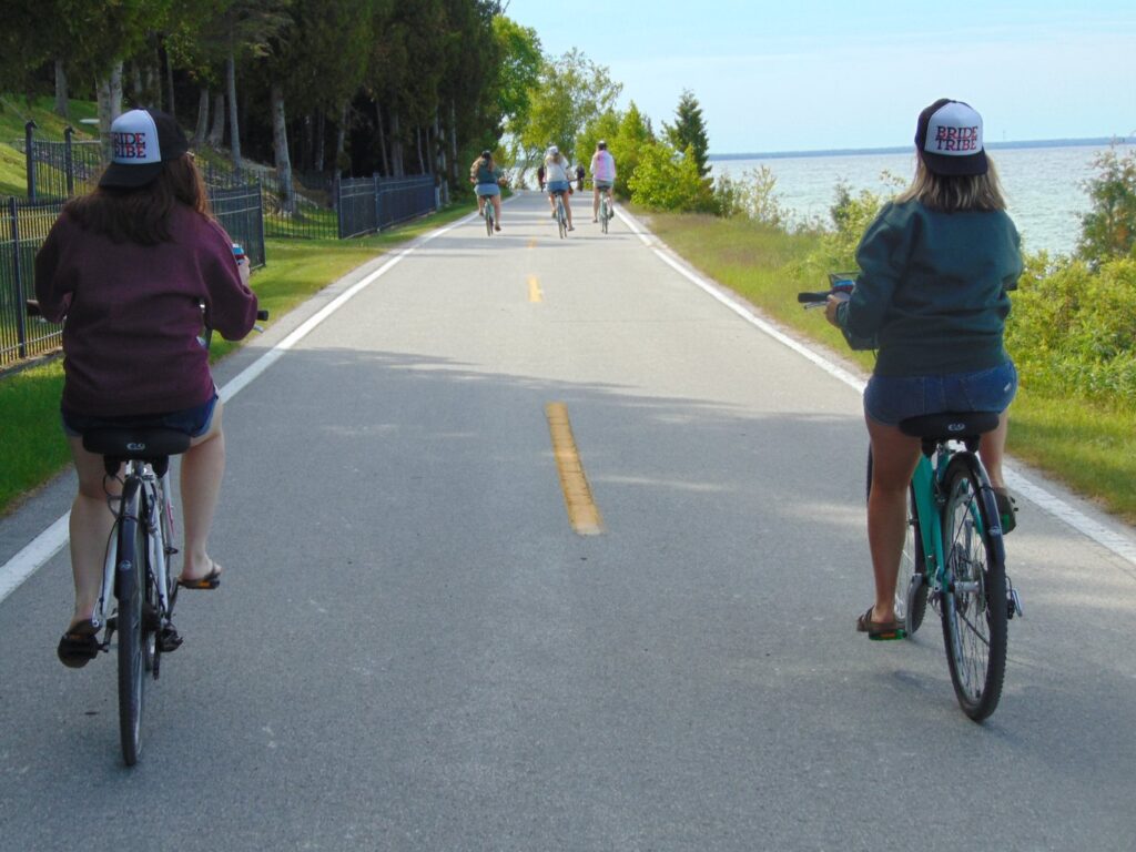 A group of people ride bicycles on a paved path beside a lake, with trees lining one side. Two people in the foreground wear matching hats and casual clothing. The scene is sunny and peaceful.