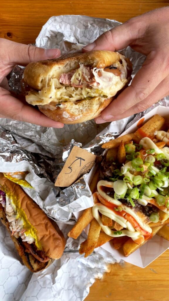Hands holding a half-eaten sandwich over a table with crumpled foil, a hot dog with mustard, and a tray of loaded fries topped with sauce and chopped green onions.