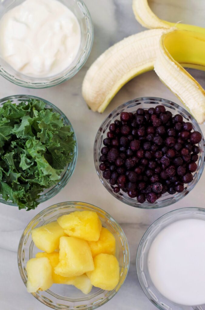 Five bowls with yogurt, kale, blueberries, pineapple chunks, and milk are arranged on a marble surface, with a peeled banana resting nearby. Ingredients appear prepared for a smoothie.