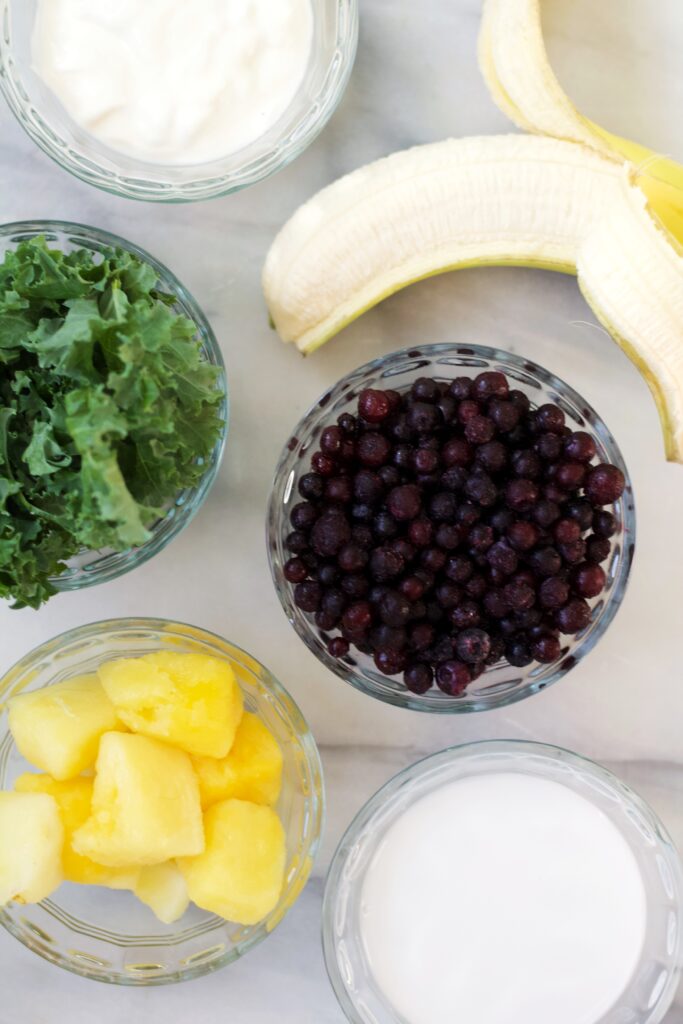 Five glass bowls containing yogurt, leafy kale, frozen blueberries, pineapple chunks, and milk, arranged around a partially peeled banana on a light surface.