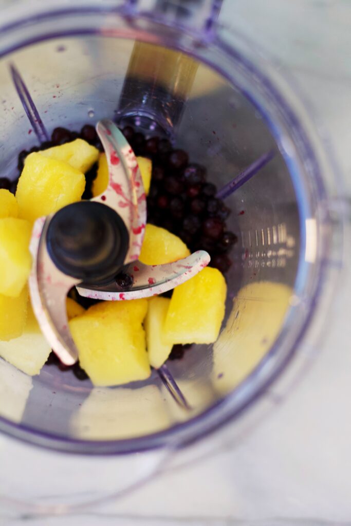 A top view of a blender with frozen pineapple chunks and blueberries inside, ready to be blended.
