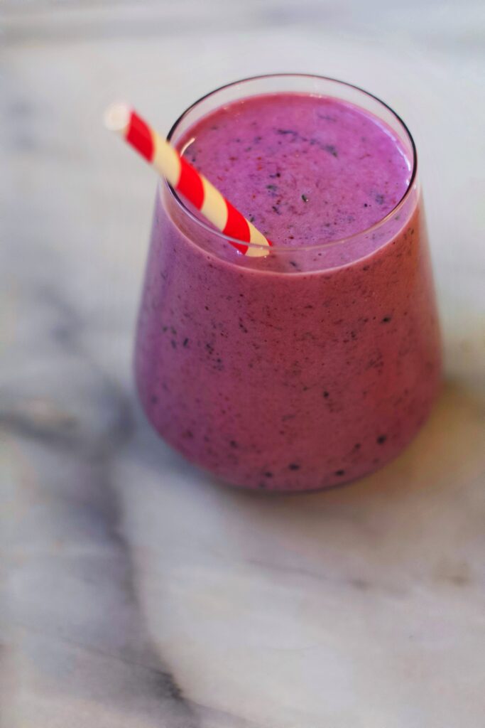 A glass filled with a thick, purple smoothie and a red-and-white striped paper straw, placed on a marble surface.