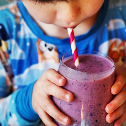 A child in colorful pajamas drinks a purple smoothie with a red and white striped straw, holding the glass with both hands.