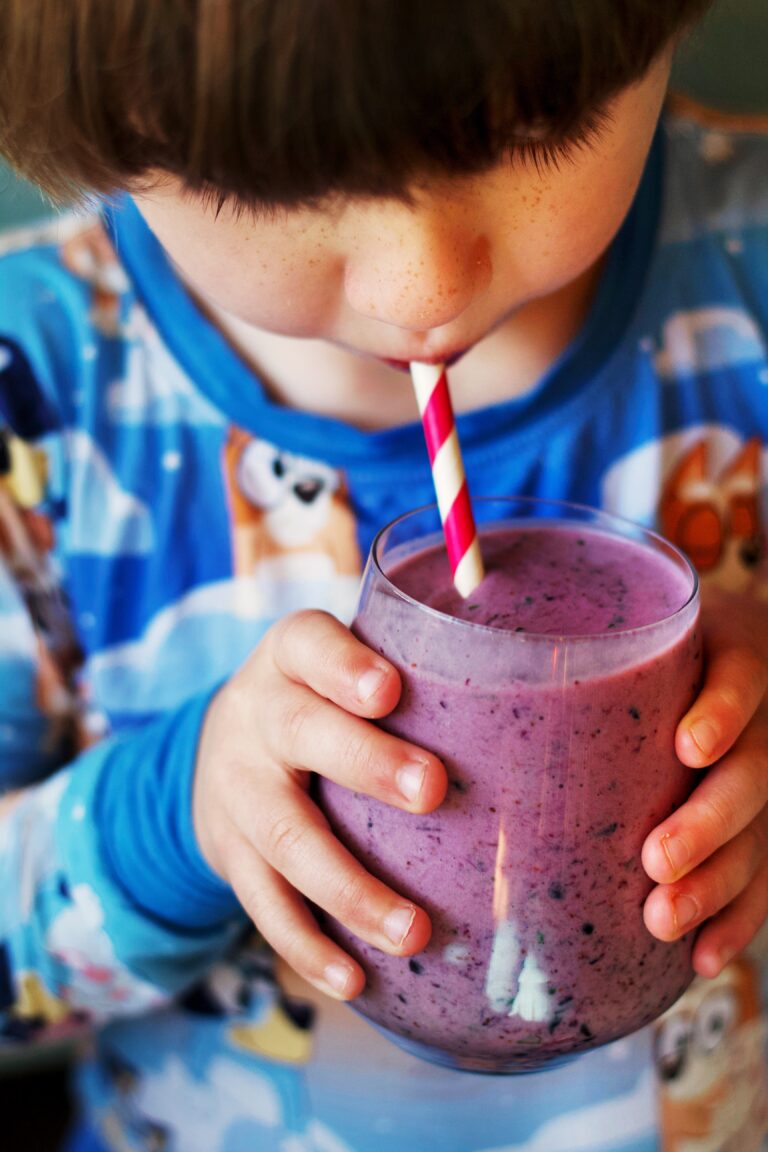A child in colorful pajamas drinks a purple smoothie with a red and white striped straw, holding the glass with both hands.