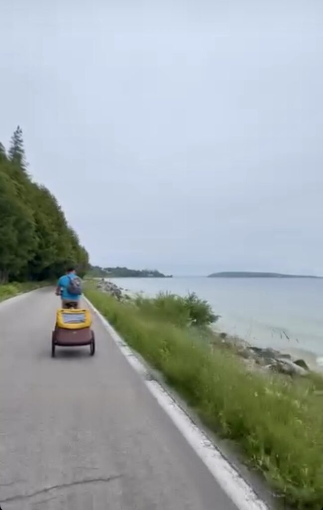 A person rides a bicycle while pulling a yellow trailer on a road beside a calm, light blue lake. Green trees line the left side, and grassy shore with rocks borders the water on the right under an overcast sky.