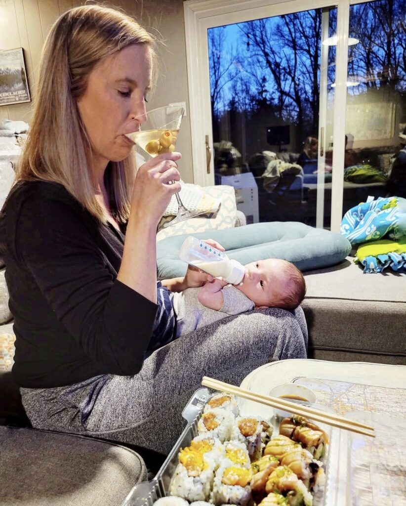 A woman sits on a couch bottle-feeding a baby while sipping a martini. A tray of sushi with chopsticks is on the table in front of her. A large window shows trees and a darkening sky outside.