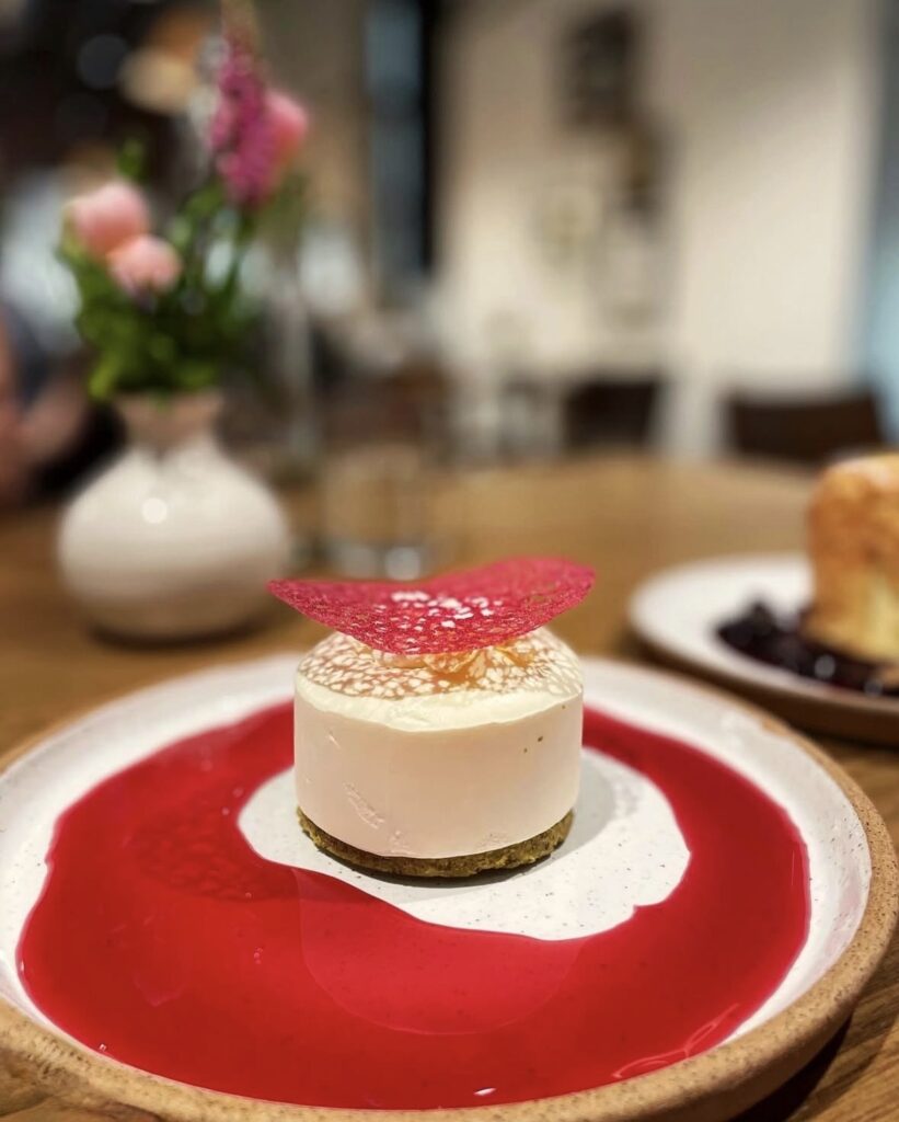 A round white mousse dessert with a thin pink sugar decoration sits on a plate surrounded by bright red sauce, with a vase of flowers blurred in the background.