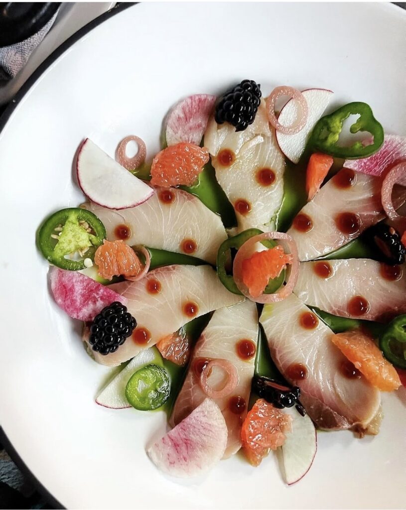 A white plate with thin slices of sashimi, garnished with radish, jalapeño, grapefruit segments, pickled onions, blackberries, and dotted with soy-based sauce, arranged in a decorative, circular pattern.