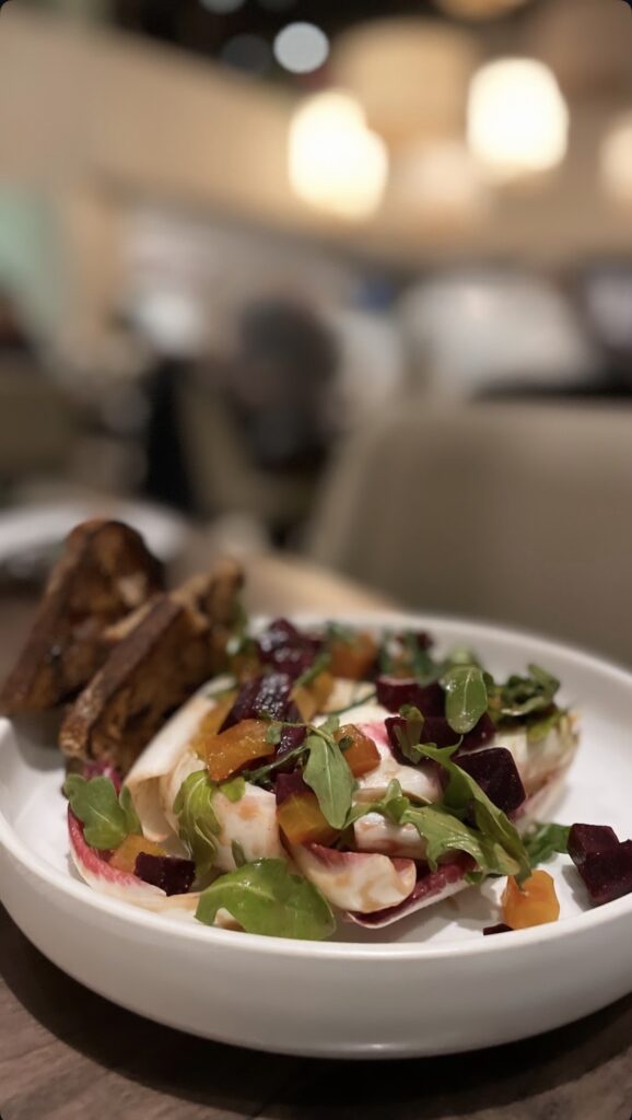 A white plate with a colorful salad of beets, greens, and orange pieces, garnished with creamy white sauce, served alongside several slices of toasted bread on a restaurant table.