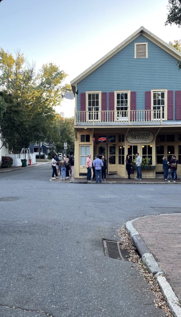 A group of people stands in line outside a two-story blue building with red shutters and a balcony on a street corner, under clear skies and surrounded by trees.