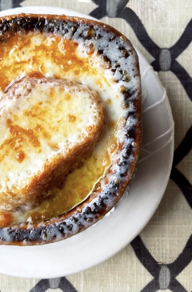 Close-up of a bowl of French onion soup topped with melted, browned cheese and a slice of toasted bread, served on a white plate with a patterned tablecloth underneath.