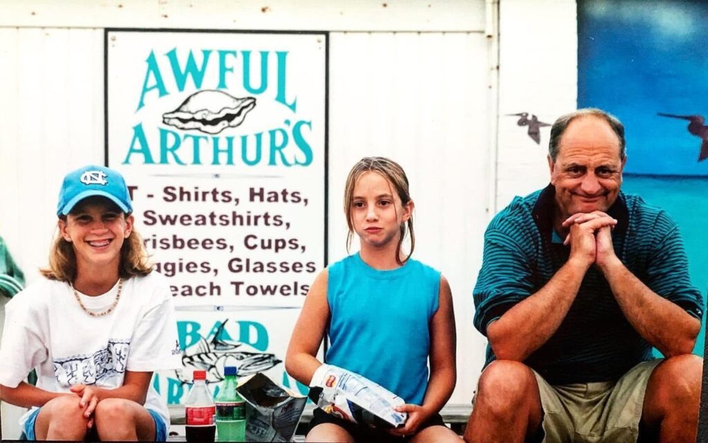 Three people sit in front of a sign for Awful Arthurs. Two girls sit on the left, one smiling and wearing a blue cap, the other holding a bag of chips. An older man sits to their right, smiling with his hands clasped under his chin.