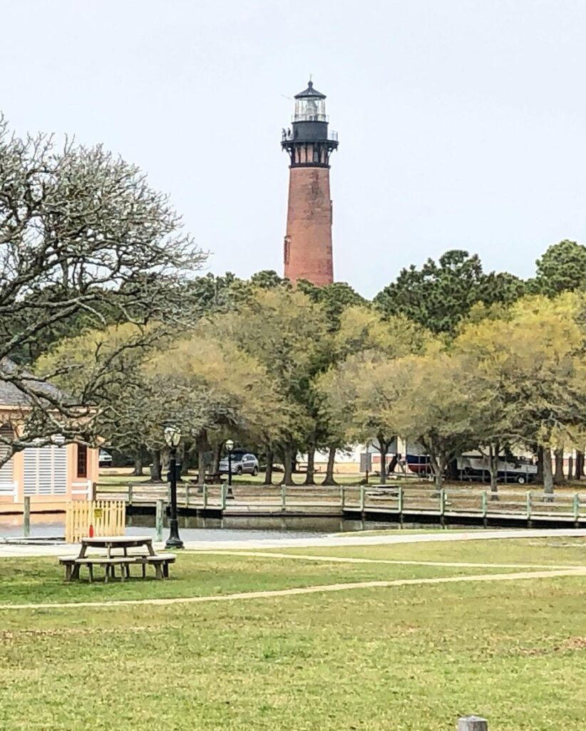 A tall brick lighthouse rises above green trees and a grassy park with picnic tables, benches, and a wooden fence in the foreground under a cloudy sky.