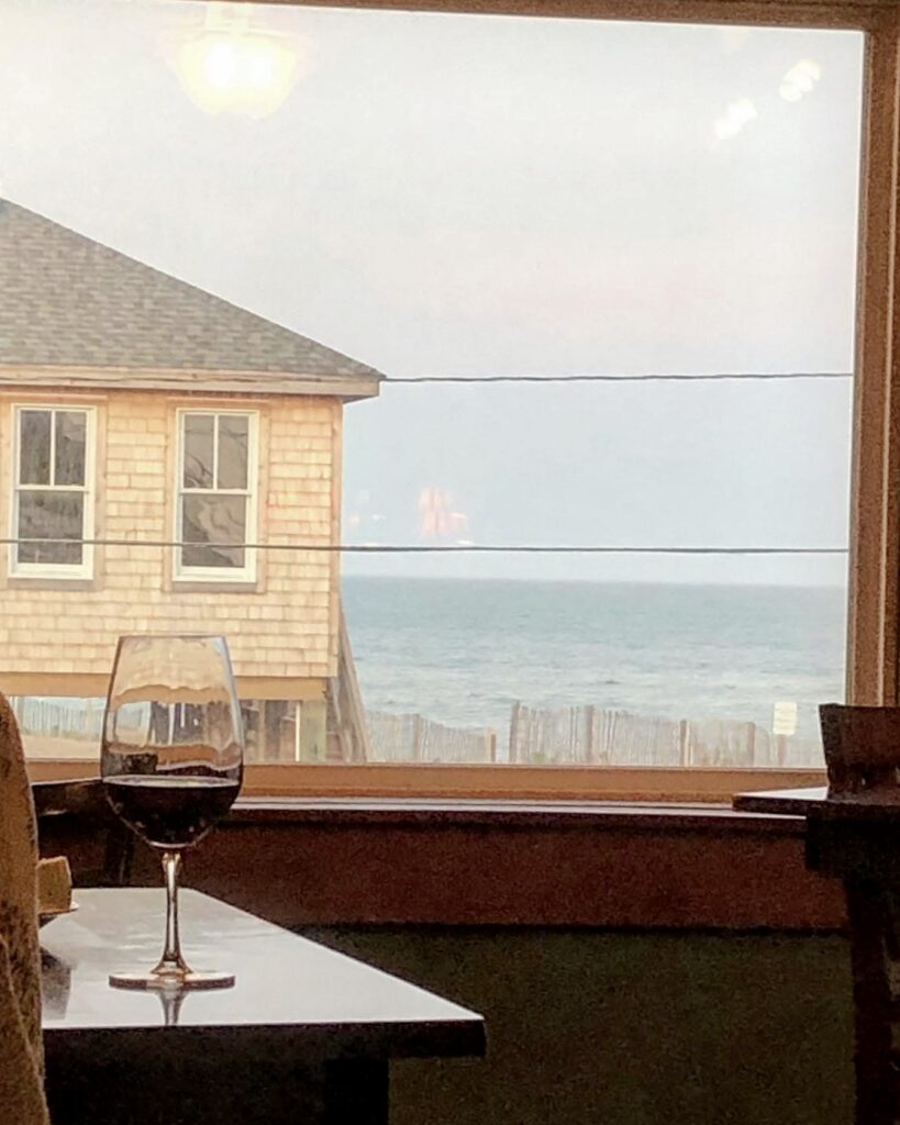A glass of red wine sits on a table by a window with an ocean view, a wooden house to the left, and waves visible under a cloudy sky.