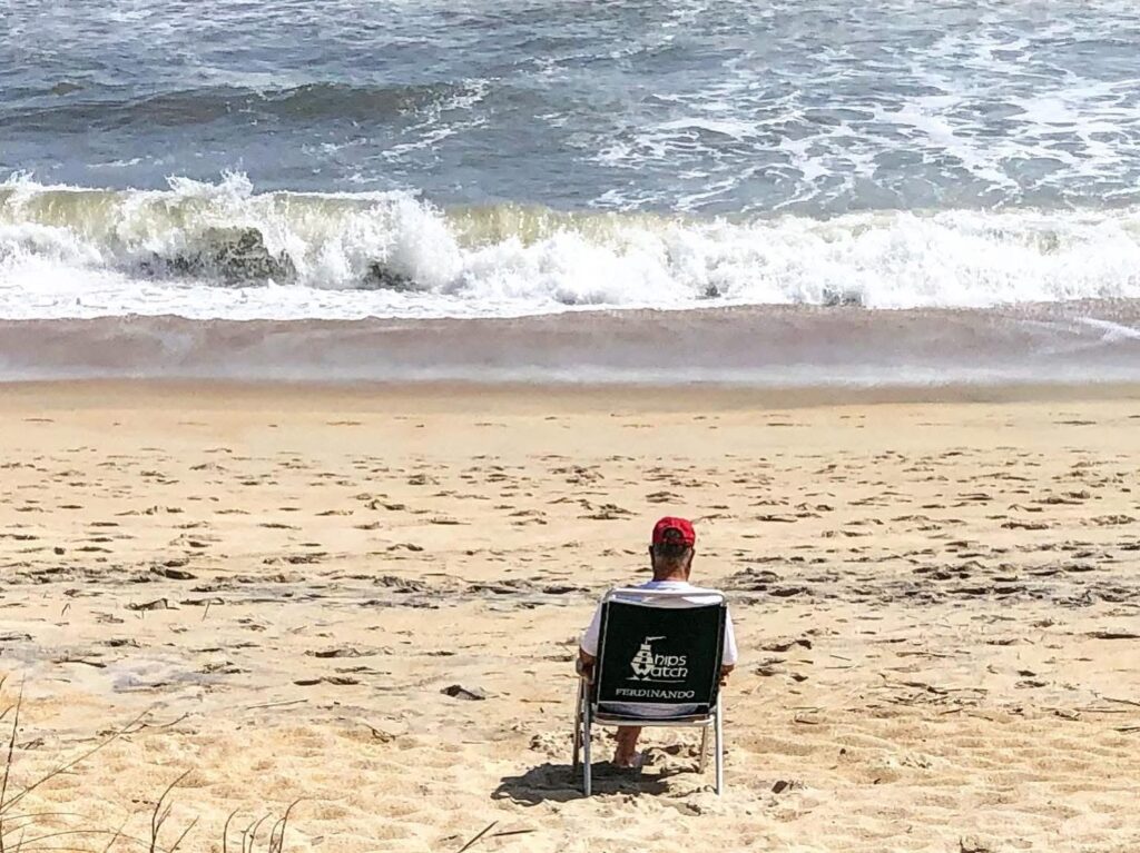 A person wearing a red cap sits alone in a folding chair facing the ocean on a sandy beach, with waves crashing onto the shore in the background.
