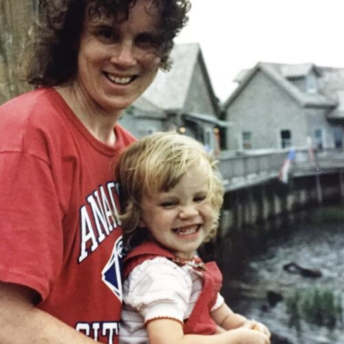 A smiling adult with curly hair and a red t-shirt hugs a grinning toddler in red overalls by a waterfront with wooden buildings in the background.