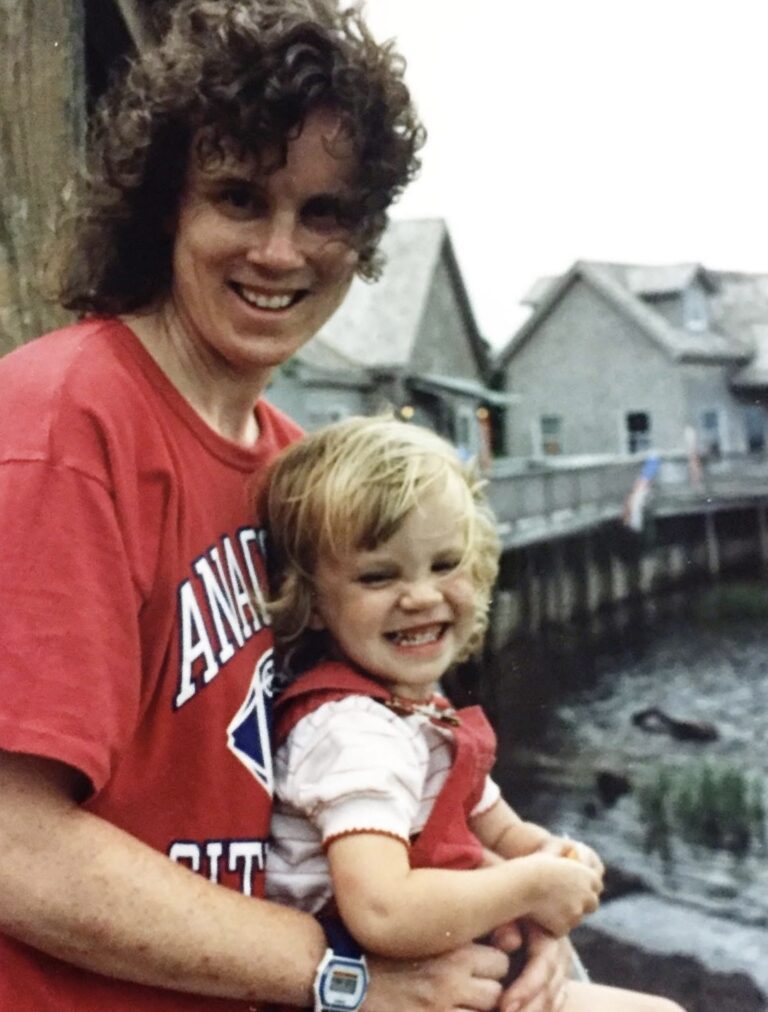 A smiling adult with curly hair and a red t-shirt hugs a grinning toddler in red overalls by a waterfront with wooden buildings in the background.