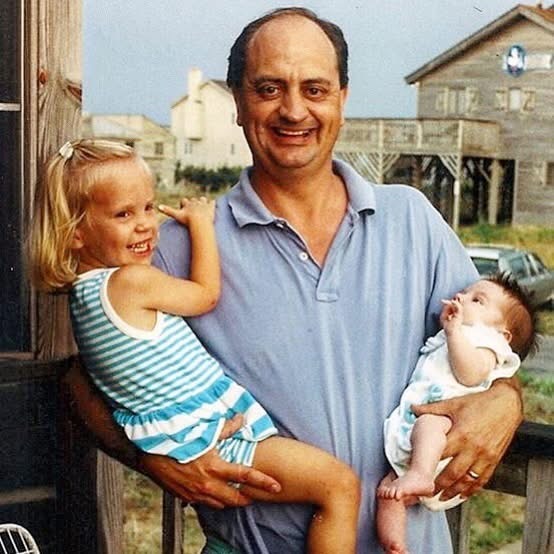 A smiling man in a blue polo shirt holds a young girl in a striped outfit and a baby in his arms, standing outdoors near wooden buildings on a sunny day.