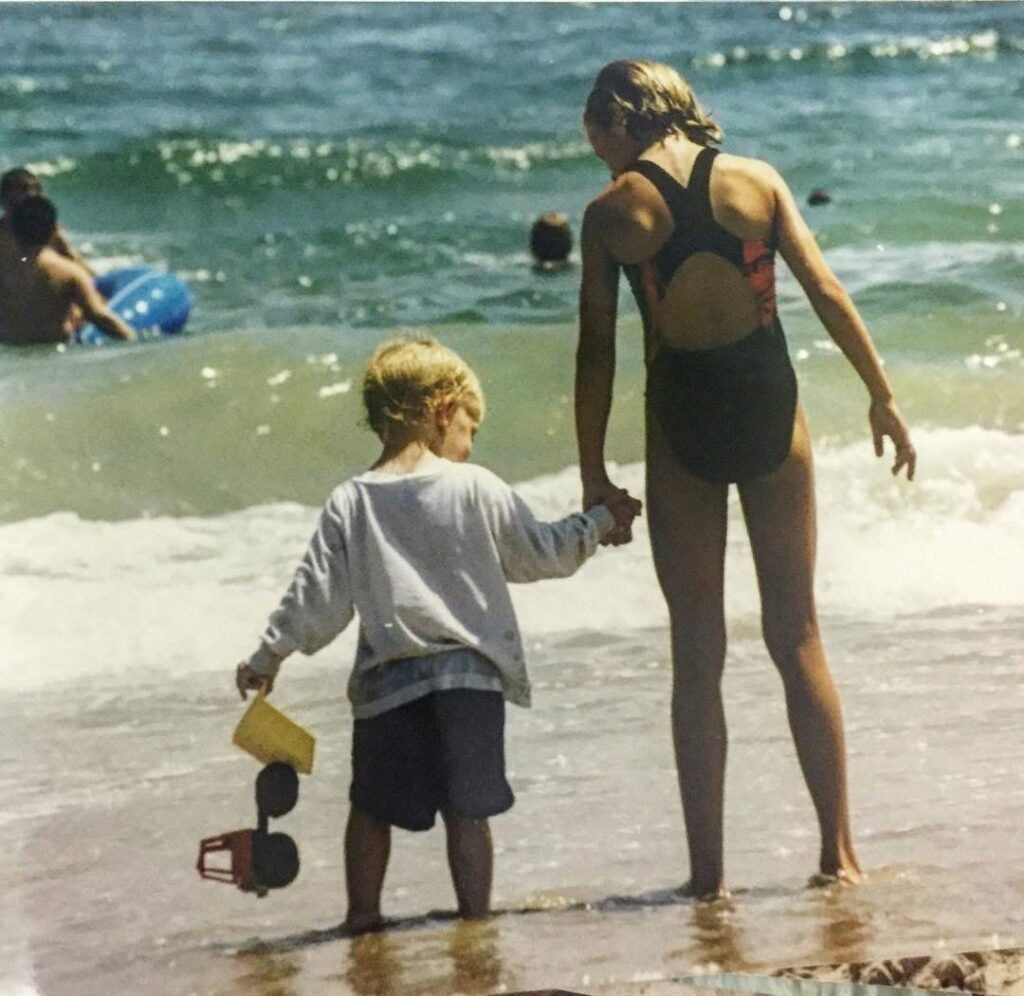 A young girl in a swimsuit holds hands with a small boy holding a toy truck as they stand at the edge of the ocean, facing the water, with other people swimming in the background.