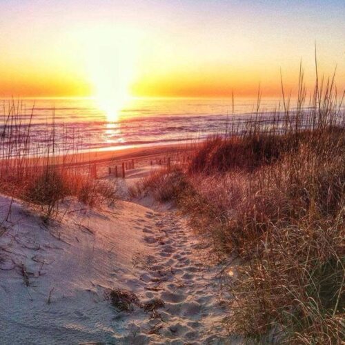 A sandy path leads through tall beach grass toward the ocean, where the sun is setting, casting a warm golden glow over the waves and sky.
