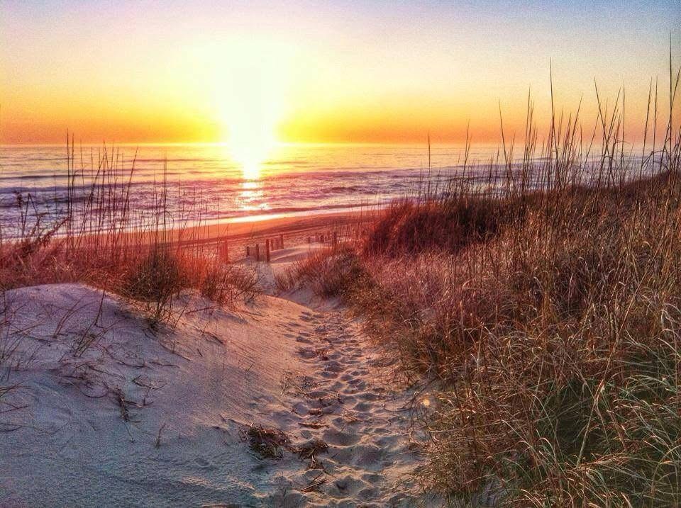 A sandy path leads through tall beach grass toward the ocean, where the sun is setting, casting a warm golden glow over the waves and sky.