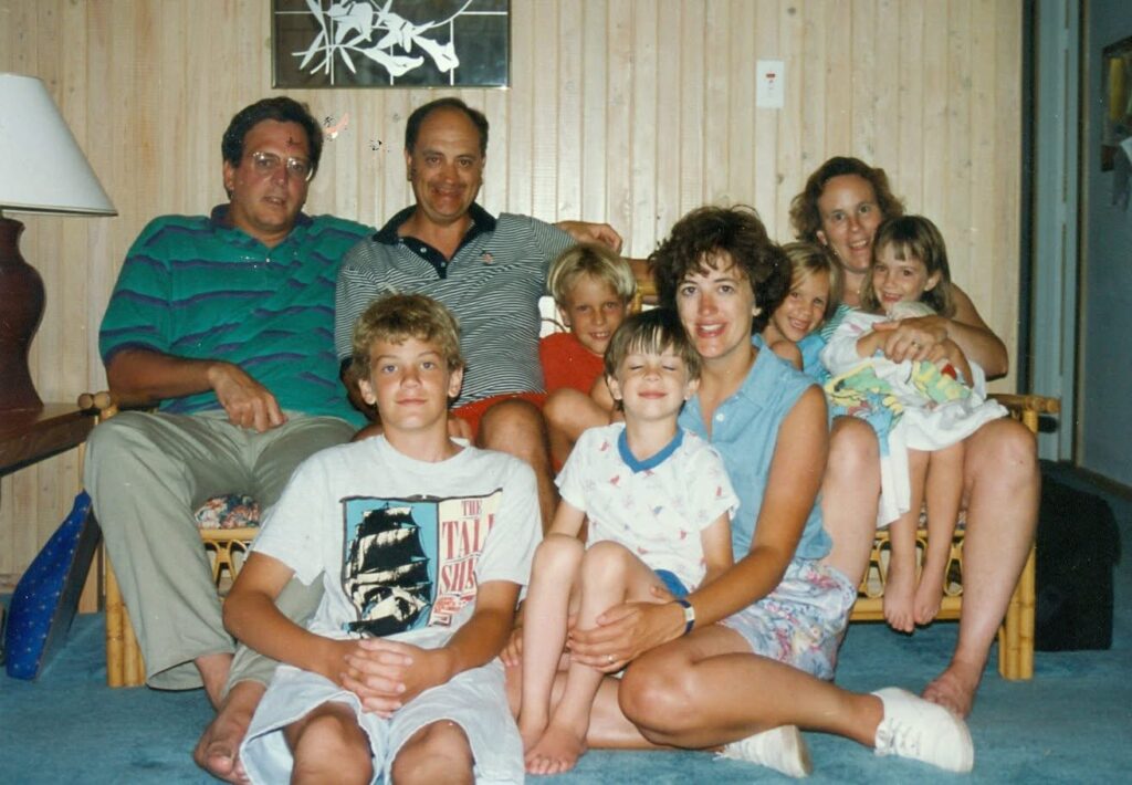 A group of nine people, including adults and children, sit closely together on a couch and on the floor in a cozy, wood-paneled room, smiling at the camera. A lamp and framed art are visible in the background.