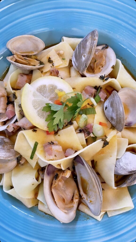 Wide pasta noodles topped with clams, diced ham, colorful diced vegetables, and herbs. A lemon slice and parsley garnish rest in the center, all served on a bright blue plate.