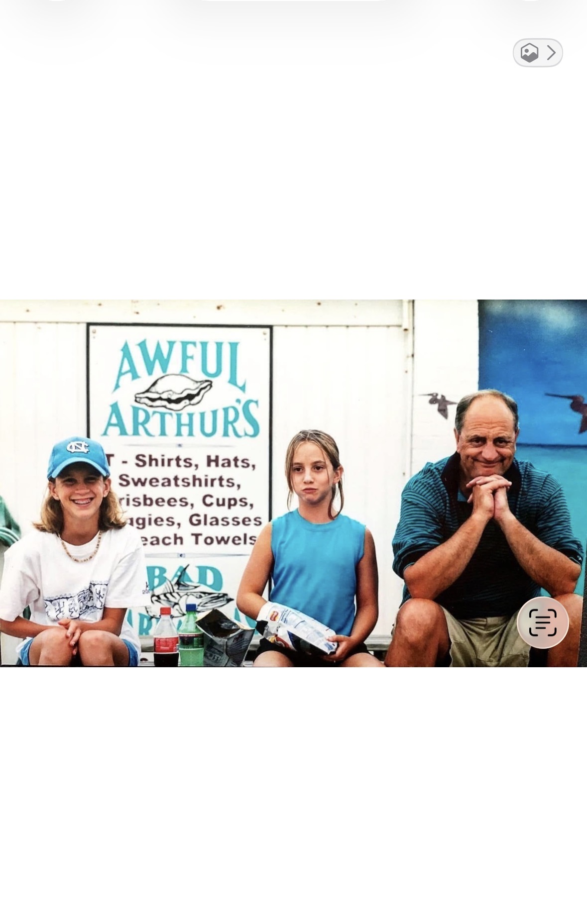 Three people sit on a bench outside Awful Arthur’s; a smiling woman in a blue cap, a girl in a blue shirt holding chips, and an older man with glasses seated thoughtfully, with a store sign behind them.