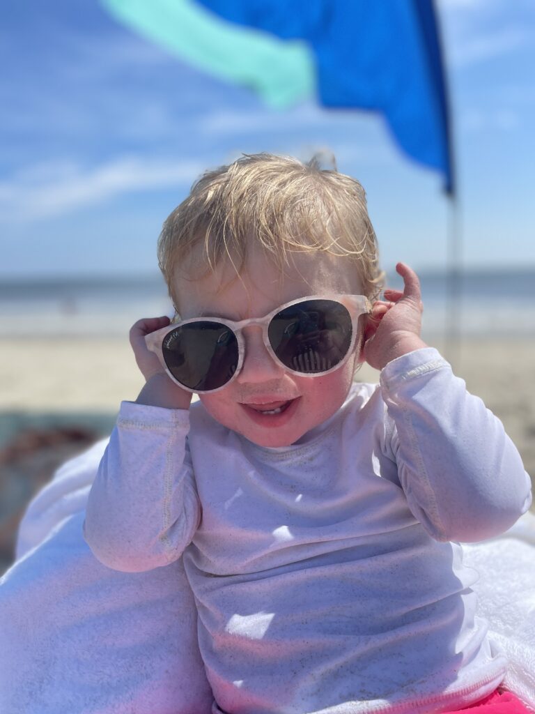 A young child wearing oversized sunglasses and a white long-sleeve shirt smiles while sitting on the beach, with sand, the ocean, and a blue flag visible in the background.