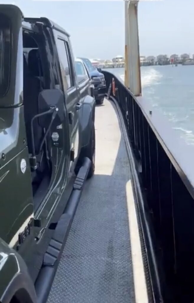 Cars are lined up on the deck of a ferry, with the ocean visible to the right and distant buildings along the horizon under a clear sky.