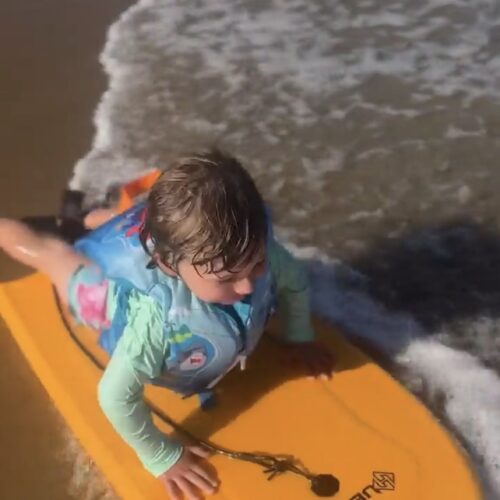 A young child wearing a blue life jacket lies on a yellow boogie board at the edge of the shore, with a small wave washing up on the sand beside them.